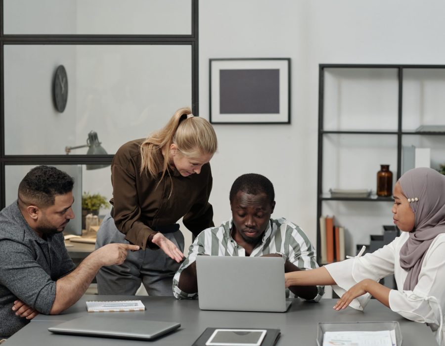 Group of intercultural displeased employees bullying African American male colleague sitting by workplace in front of laptop